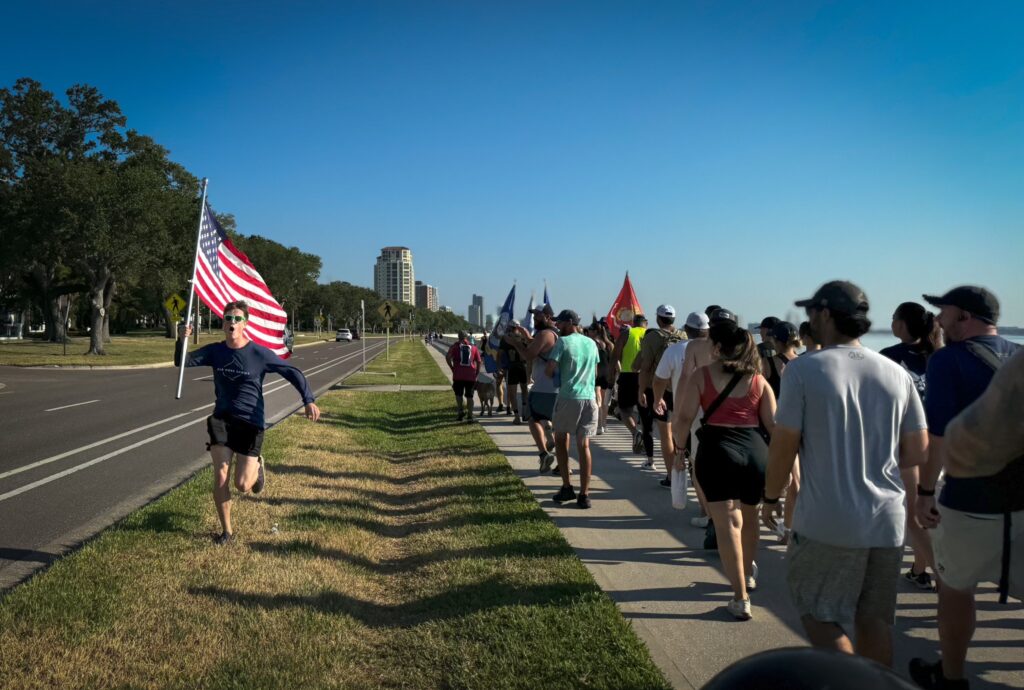 Power Tampa employees march along the water while one employee runs with an american flag for their annual Memorial Day Ruck March.