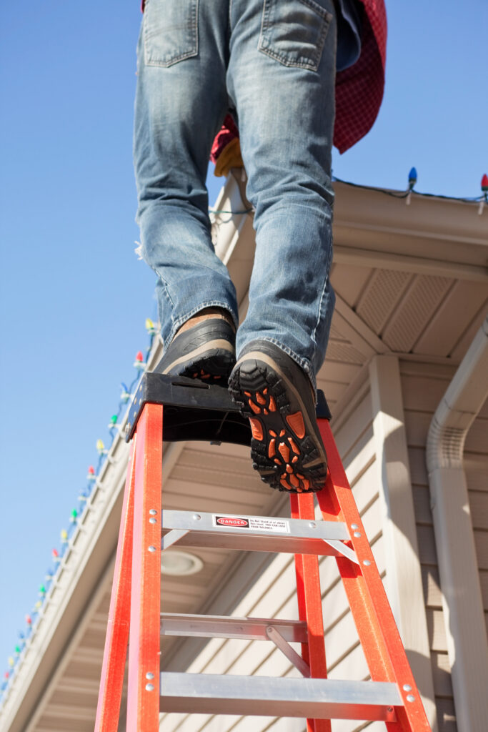 A man installing Christmas lights on the roof of his house, dangerously steps onto the top of a tall ladder