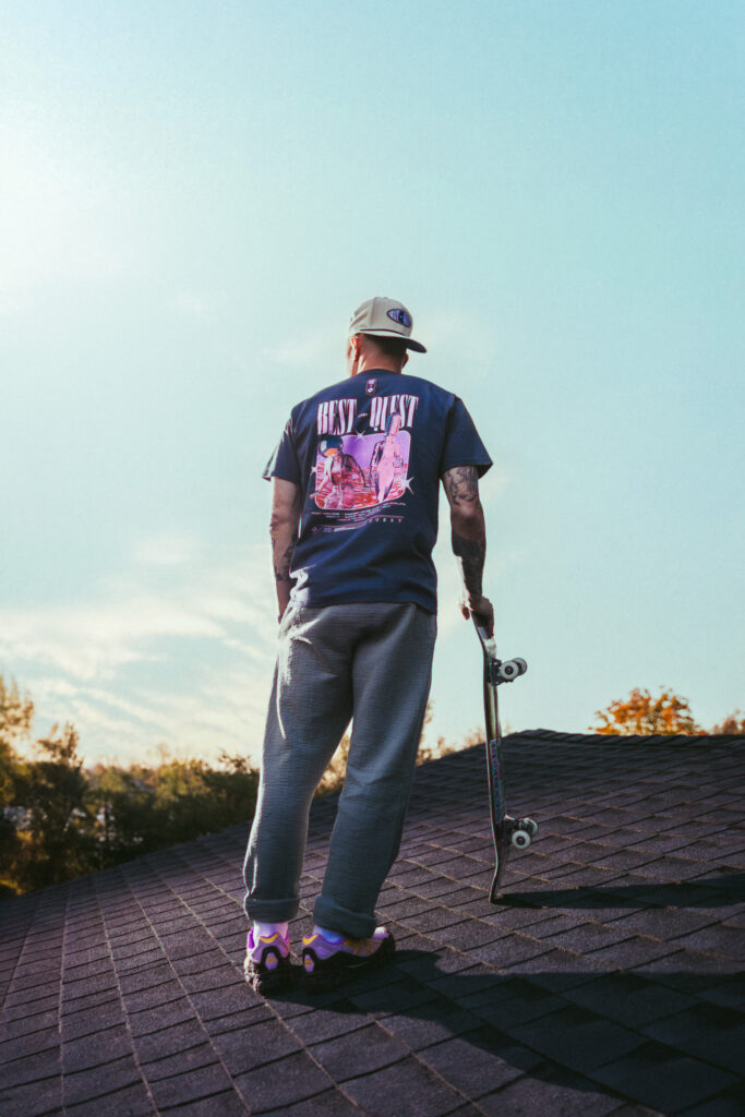 Jimmy Gorecki poses on a roof with a skateboard showing the back of the POWER x Standard Quest lineup t shirt.